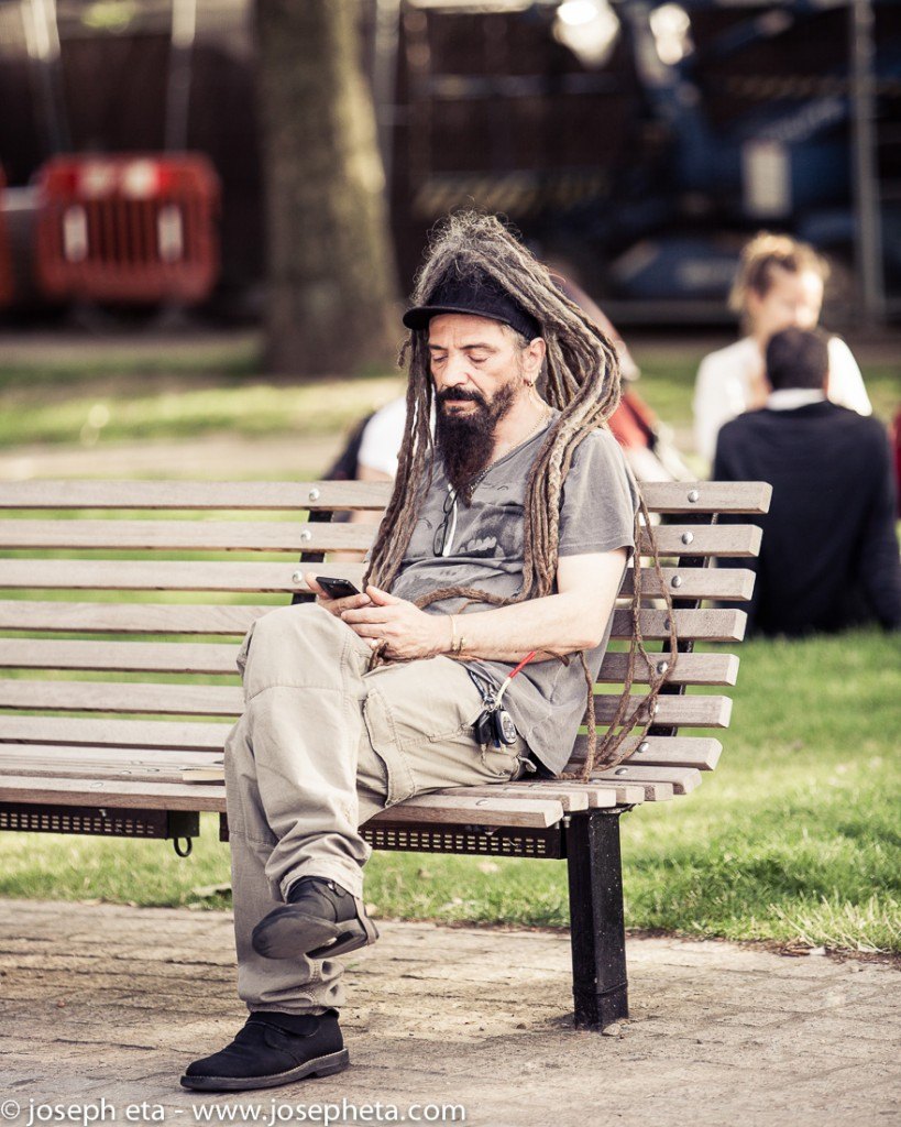 street photography of a man with very long dred-locks sitting at the South Bank in London and reading messages on his phone