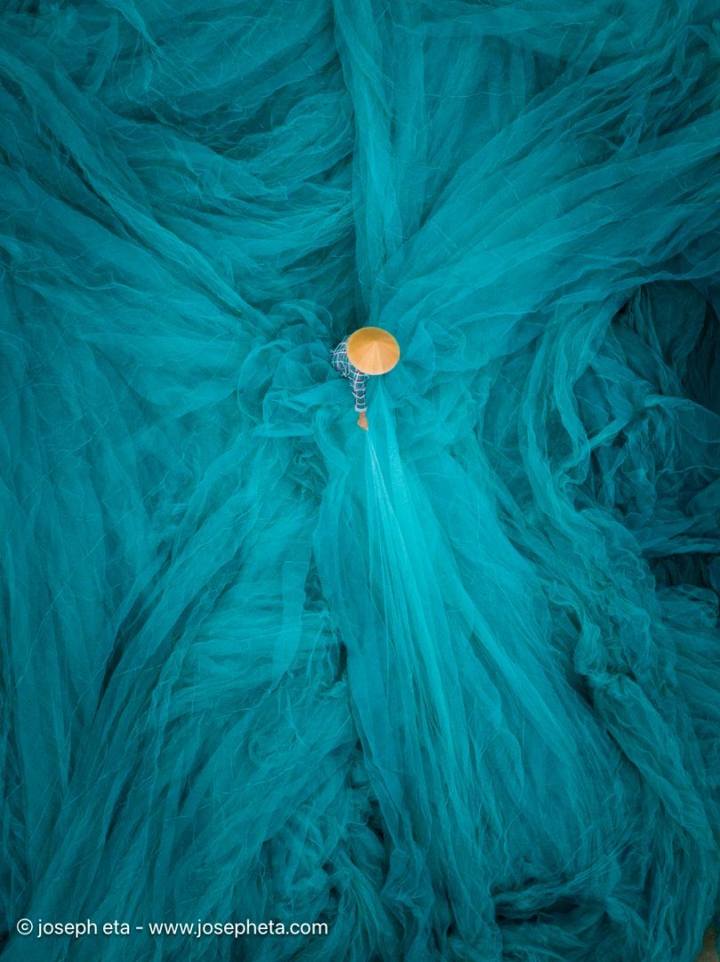 Abstract aerial photograph of a fisherman casting a net in teal water, creating a geometric circular pattern. Fine art print showcasing human craftsmanship and aquatic textures for boutique interiors.