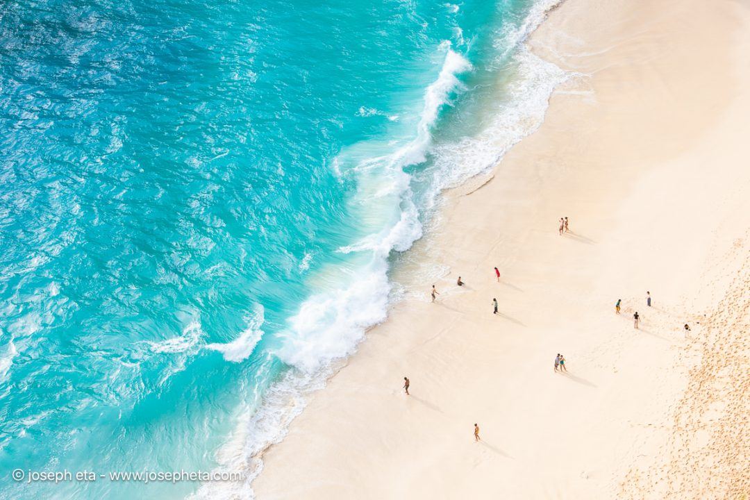 High-key aerial beach photography featuring bright turquoise water, pale white sand, and tiny figures on the shoreline. An optimistic, social art piece representing the 2026 Transformative Teal trend.